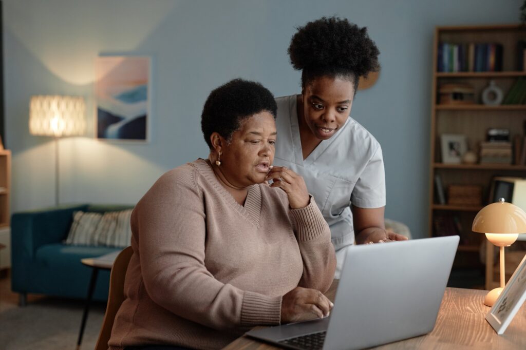 Black Female Caregiver Assisting Senior Woman with Online Payment on Laptop at Home